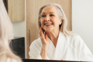 Happy aged female touching her face in front of a mirror. Woman in bathrobe admires her reflection.