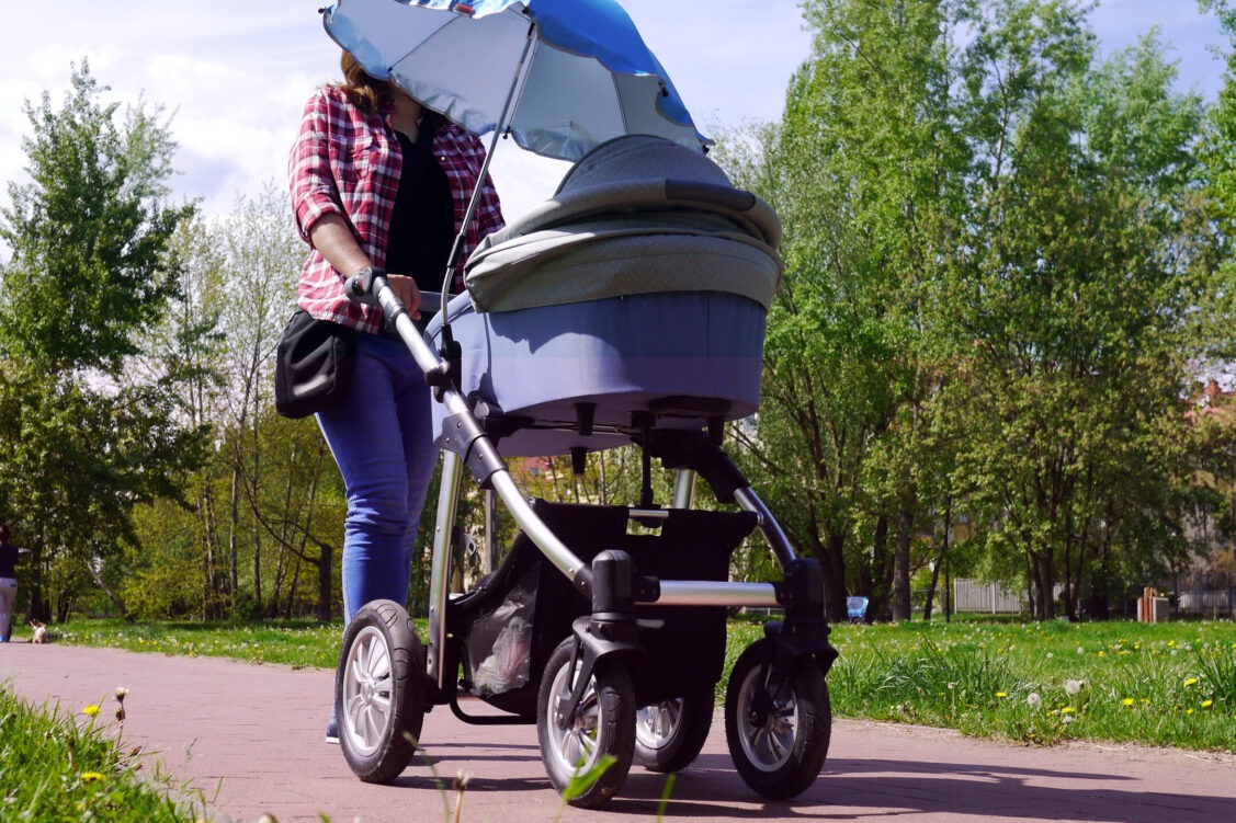 Mama z nowo narodzonym dzieckiem w wózku. Kobieta spaceruje w parku blisko lasu. Dziecko na świeżym powietrzu, dbanie o zdrowie dziecka. Noworodek śpi na spacerze. Urlop macierzyński. Słoneczny dzień.