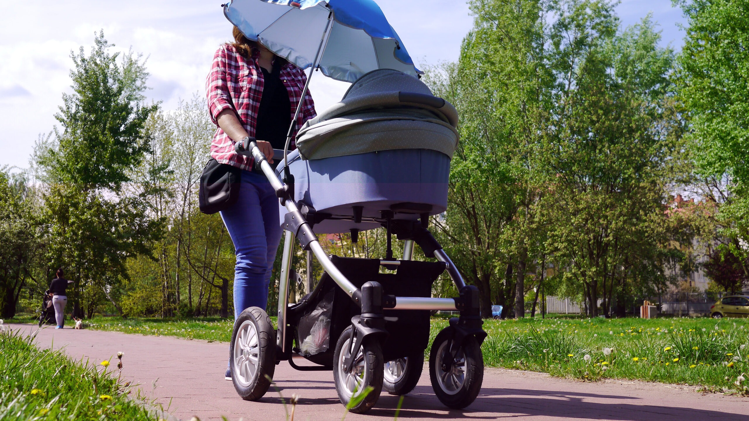 Mama z nowo narodzonym dzieckiem w wózku. Kobieta spaceruje w parku blisko lasu. Dziecko na świeżym powietrzu, dbanie o zdrowie dziecka. Noworodek śpi na spacerze. Urlop macierzyński. Słoneczny dzień.